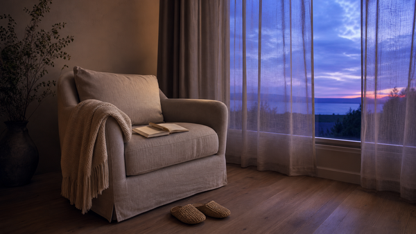A single armchair beside a soft-lit window at dusk, with a folded throw blanket and a small open notebook — the quiet starting point of a five-minute evening ritual.